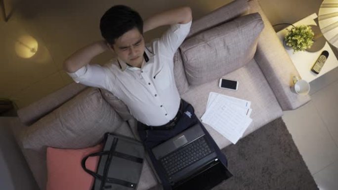 从上面看，压力很大的亚洲男性员工在家里客厅的笔记本电脑上工作到很晚时，向后仰着头，向上看。