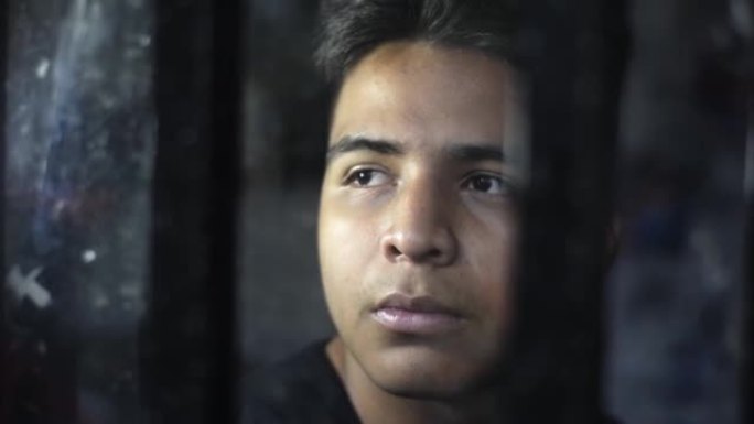 一个年轻的拉丁男人的脸的特写镜头，透过窗户非常沉思。监狱里的人，在黑暗的房间里