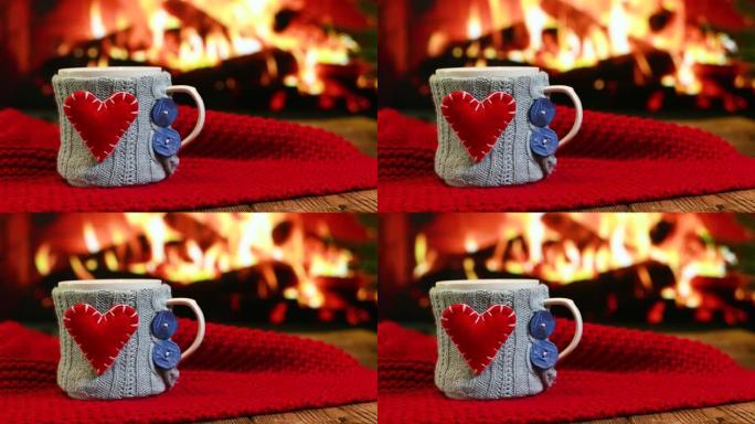 在燃烧的壁炉和红色格子的背景下，在木制桌子上放着带有红色心形的蓝色针织罩子的热饮杯子