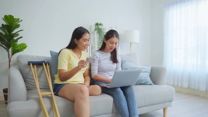 亚洲女性截肢者与朋友在客厅使用笔记本电脑。迷人的年轻无腿女孩和室友坐在沙发上，花空闲时间在家里一起网