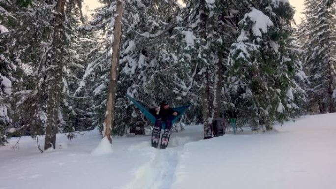 徒步旅行者带着雪鞋喝咖啡，躺在冬季森林的吊床上