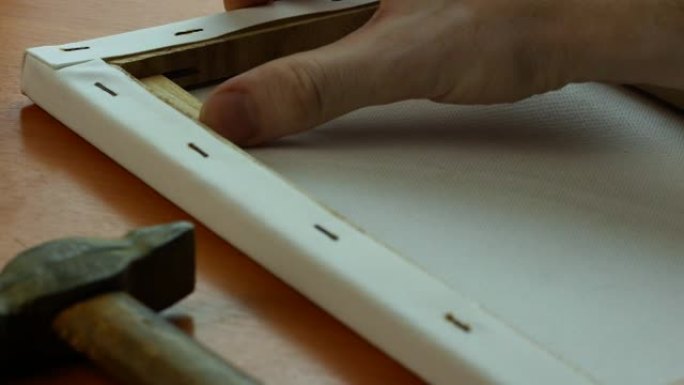 一位工匠用锤子敲打木制垫片来拉伸画布的特写镜头，制作相框。艺术家为这幅画准备画布。选择性聚焦