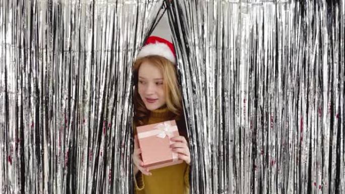 有着红头发和圣诞老人帽子的年轻女孩带着礼物从闪亮的窗帘上望去，环顾四周