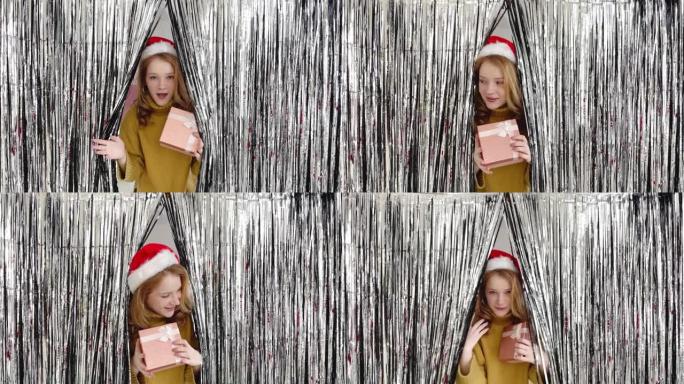 有着红头发和圣诞老人帽子的年轻女孩带着礼物从闪亮的窗帘上望去，环顾四周