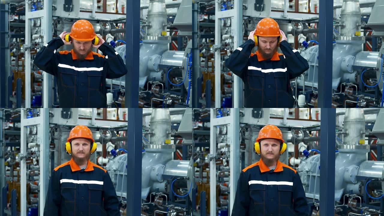 工作场所的大胡子男性工业机械师。戴着头盔的工人在靠近设备时戴上耳机，在工作中会产生很大的噪音。在压缩