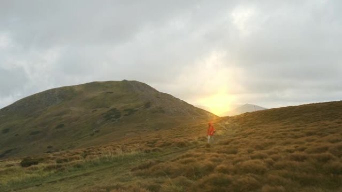 日落在山中，一个旅游女孩沿着一条绿色的小路散步，背景是山后落下的橙色太阳。宽镜头，春夏，喀尔巴阡山脉
