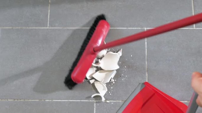 一个女人的特写镜头，用红色的塑料勺子从地板上捡起一个破碎的白色陶瓷杯的碎片。打碎的盘子从地板上移开，