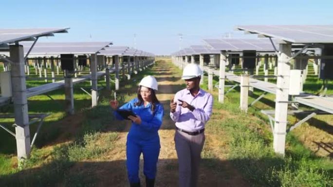 非洲投资者和亚洲中年工程师，穿着蓝色防护服，安全眼镜，头盔，在太阳能农场散步时讨论，前高角度视图