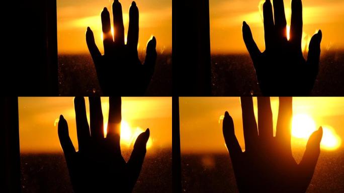 剪影女性手在玻璃窗上，面对日落，手指之间有阳光