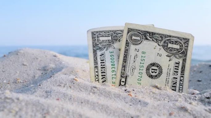 阳光明媚的夏日，海蓝色天空背景下的沙子里有两张旧的美元钞票。肮脏的美元特写。钱是从沙子里长出来的。抽