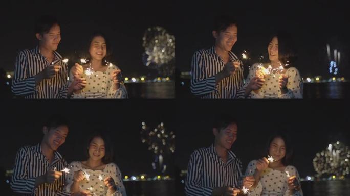 这对幸福的夫妇手持烟火灯，晚上在海滩边的烟花节上欣赏烟花汇演。