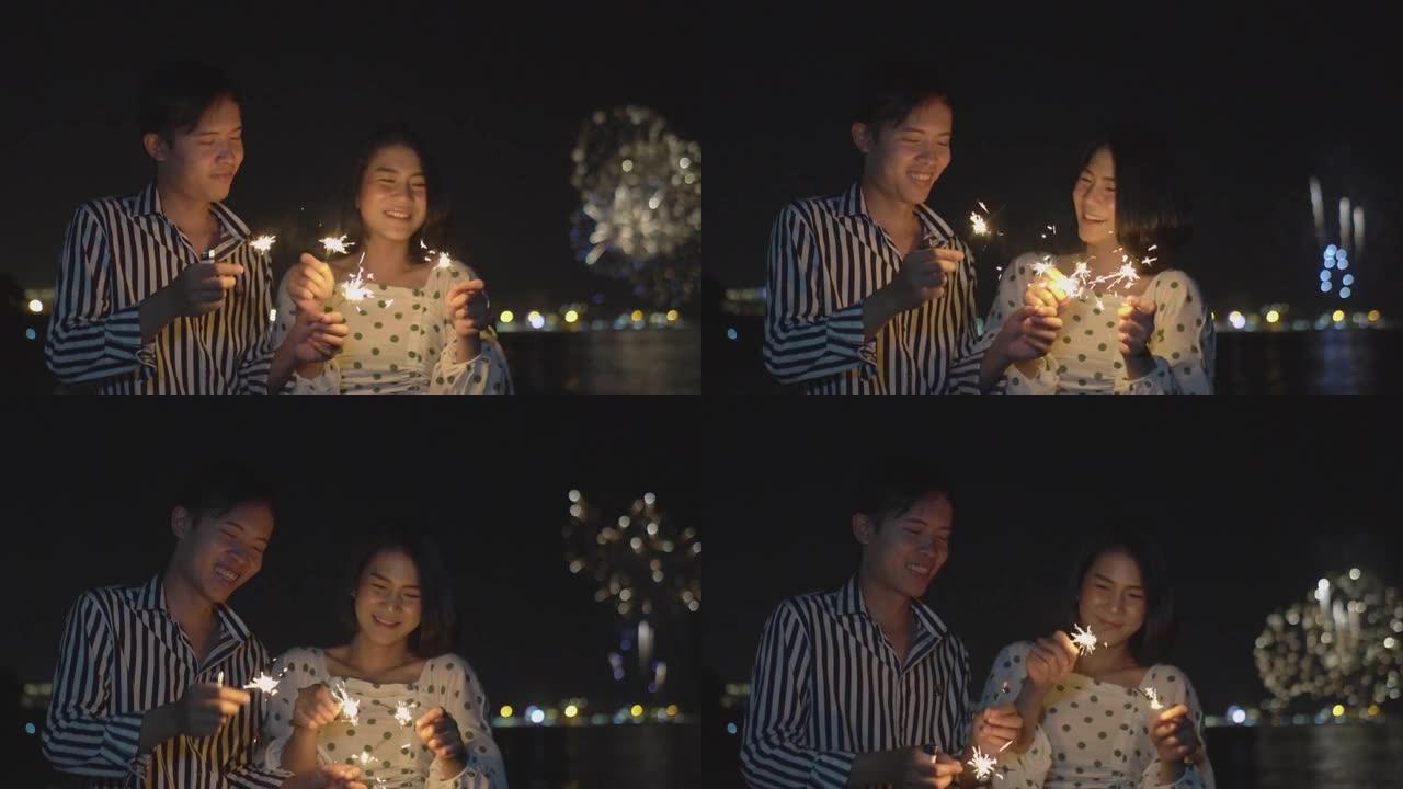 这对幸福的夫妇手持烟火灯，晚上在海滩边的烟花节上欣赏烟花汇演。