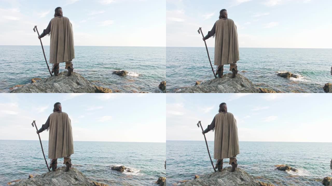 一个拿着斧头的中世纪骑士站在石头上，望向大海。后视图