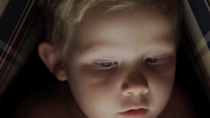 一个男孩在黑暗中的枕头小屋中的电子平板电脑上观看动画片