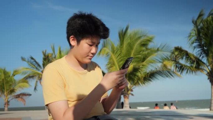 亚洲男孩在阳光明媚的日子里，在海滩上使用带有椰子树背景的智能手机，生活方式的概念。