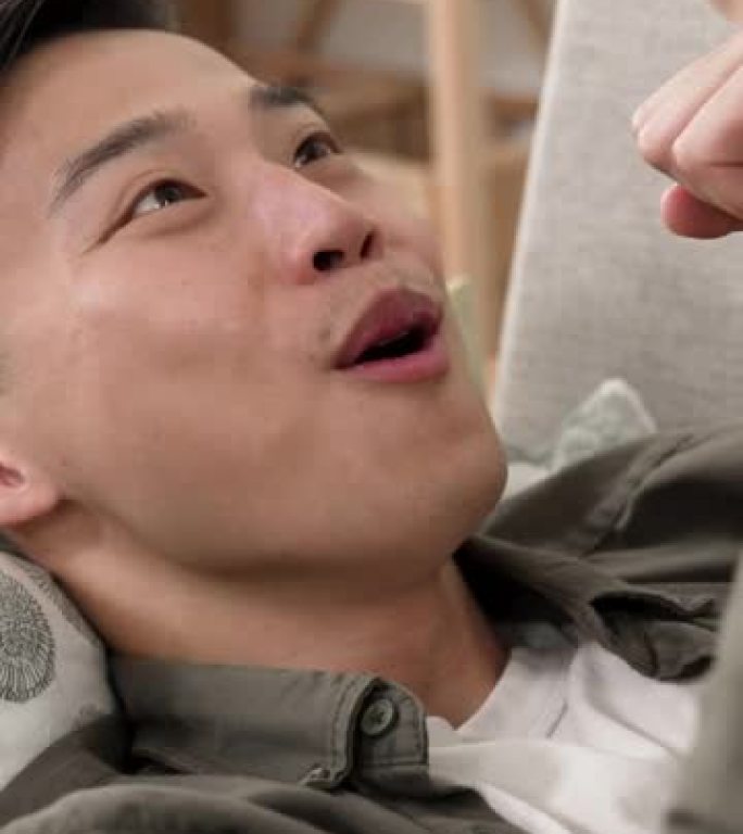 垂直视频: 一位惊讶的韩国男性在家中使用智能手机躺在沙发上时握紧拳头为在线新闻欢呼的特写镜头。