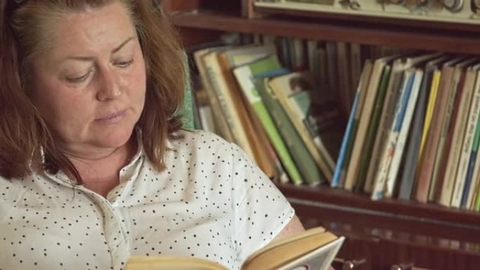 一个胖胖的中年妇女在书柜的背景下，坐在扶手椅上读一本旧书。孤独和遗弃。对自己的失望