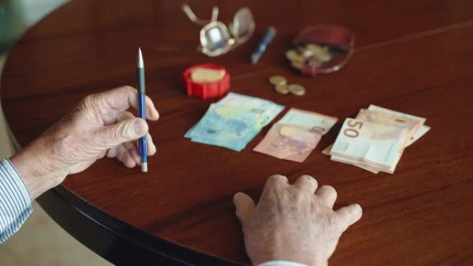 无法识别的高级男人坐在桌子旁，手里拿着写字笔，看着桌子上的纸币和硬币，退休时的男人为不同的目的分钱储