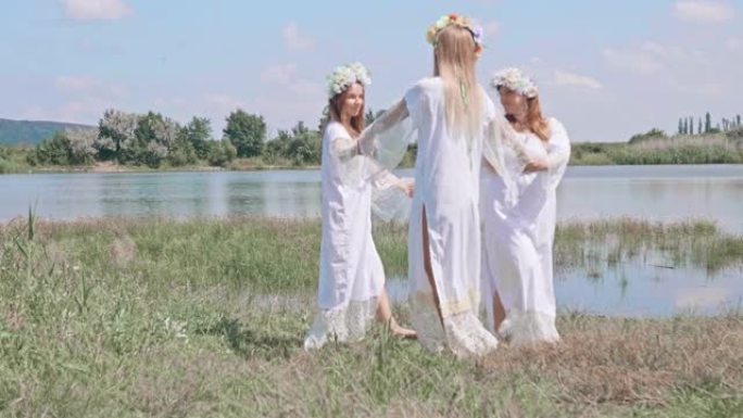 三个穿着传统白衬衫和花圈的快乐微笑的女人在天空和野生动物与湖泊的背景下跳舞。侧视图。伊凡·库帕拉的历