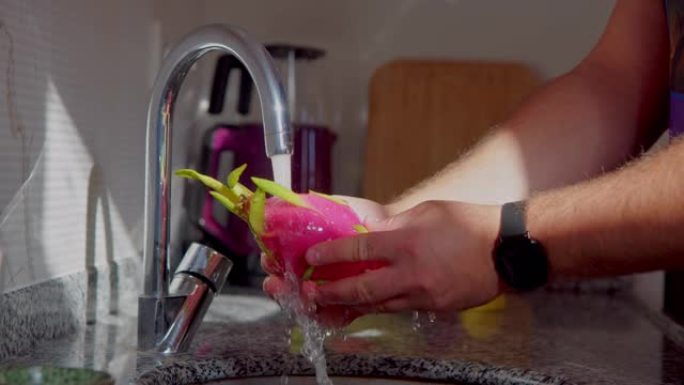 男性双手打开水龙头，洗净成熟多汁的火龙果。在流水下，明亮的粉红色pitahaya在厨房中清洗。异国水