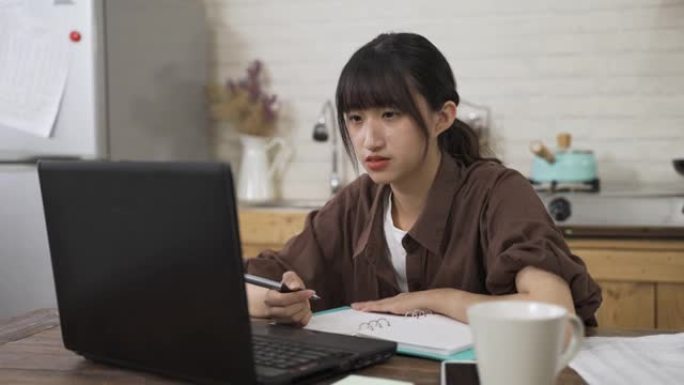 肖像亚洲女学生在家里的餐厅里通过视频聊天进行在线讨论时，一边玩笔，一边对着电脑屏幕说话。