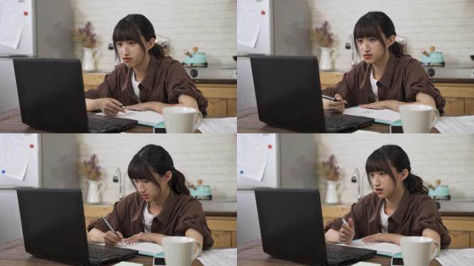 肖像亚洲女学生在家里的餐厅里通过视频聊天进行在线讨论时，一边玩笔，一边对着电脑屏幕说话。