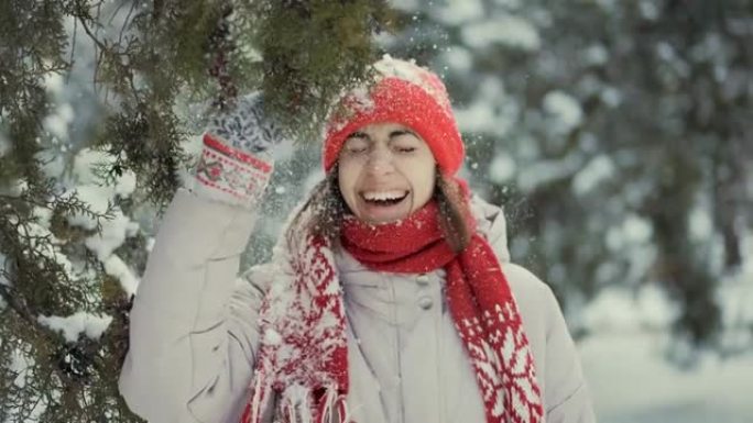4k慢动作女孩，穿着针织红领巾，无檐小便帽和手套站在公园里，摇晃杉木树枝。雪落在女孩的帽子和脸上，女