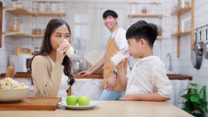 年轻的亚洲男孩在现代厨房里微笑着和母亲一起喝牛奶，母亲抚摸着他的头。父亲站在后面，看着家人。享受早餐