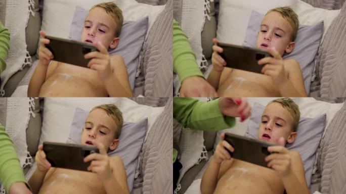 母亲在儿子的皮肤上涂上乳液，以减少水痘的瘙痒，同时他在手机上玩电子游戏