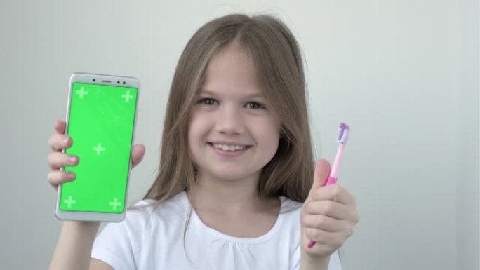 穿着白色t恤的小女孩展示绿屏手机和牙刷。色度键绿屏智能手机为牙科诊所广告设置。刷牙的孩子。保健，口腔