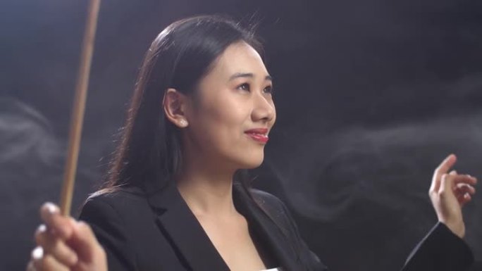 亚洲指挥家妇女的特写镜头握着警棍闭上眼睛，在迷雾的黑色工作室里展示手势