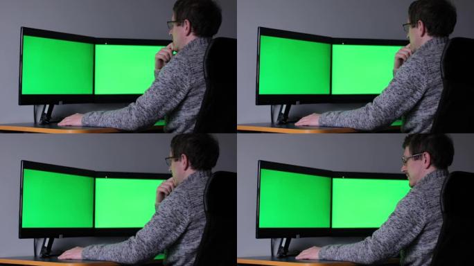 戴着眼镜和红胡子的中年专家在繁忙的创意办公室与同事一起在台式计算机上工作，带有绿屏模型。修剪胡须的男