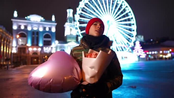十几岁的高加索男孩的肖像，一束鲜花和心形气球站在情人节之夜的户外环顾四周。充满爱心的少年在度假的城市