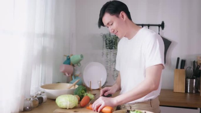 穿着白色t恤的亚洲年轻人在家里的厨房里切白菜和胡萝卜等生蔬菜。素食主义者做沙拉。