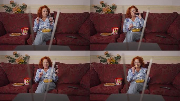 放松快乐的复古女人吃薯条喝苏打水坐在沙发上看老式电视。无忧无虑自信的高加索红发女士在家里享受休闲的前