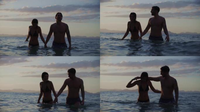 浪漫的异性恋夫妇在日落时一起在海里游泳。牵着手走过波浪