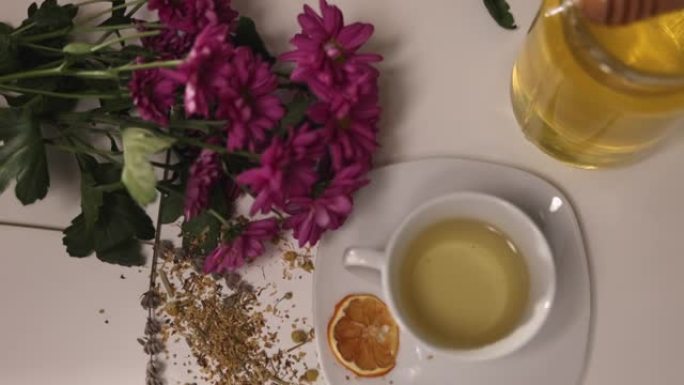 桌子上的陶瓷杯茶的高角度视图，桌子上放着草药，蜂蜜罐，鲜花