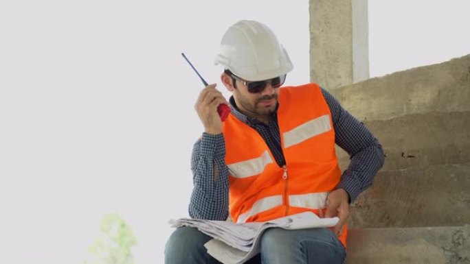 工头戴安全帽，手持图纸，通过智能手机与施工现场的工人进行检查或控制和沟通。承建商在物业发展行业使用无