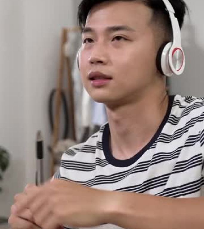 垂直视频: 分心的亚洲年轻人从家里学习，正在玩笔像鼓棒，同时在卧室的办公桌上听耳机上的流行歌曲。