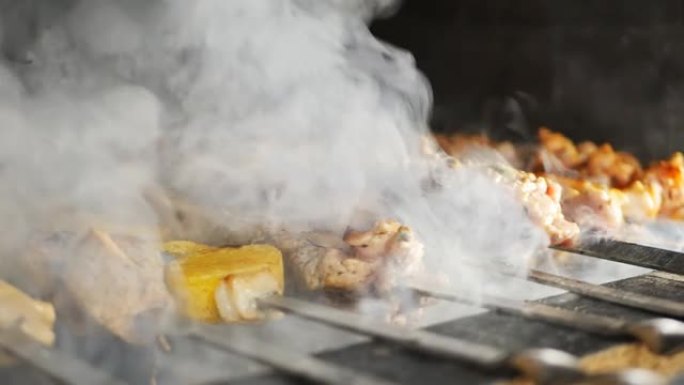 在烘烤过程中，浓烟在餐厅厨房的烤架上的金属串上串起了带有金色土豆块的美味肉类烧烤。