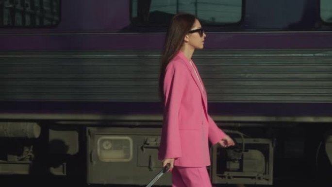 一位穿着粉红色西装，带着手提箱的年轻女子在火车附近的平台上行走，特写镜头跟随拍摄