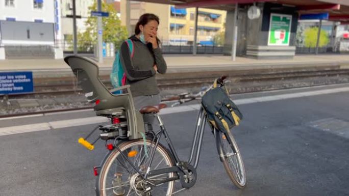 疲倦的女人站在站台上打哈欠，等待火车带着自行车到达。早上通勤的人