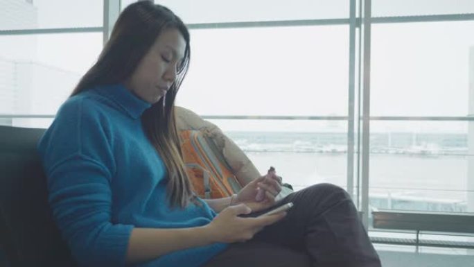 东南亚年轻女性旅行者，手持手机坐在慕尼黑国际机场的玻璃窗旁，滚动智能手机应用程序，检查航班