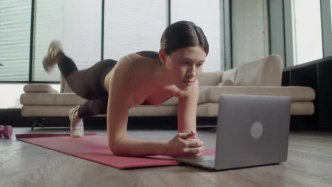 穿着运动服的亚洲运动女性在家锻炼。女性健身在打开的笔记本电脑前摇晃她在瑜伽垫上的按压，重复专业健身教