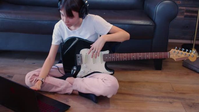 戴着耳机的吉他的少女坐在笔记本电脑前的地板上。