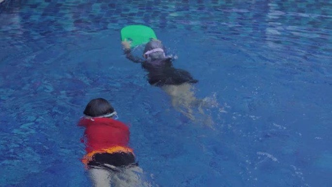 4K，两个亚洲孩子在游泳池里游泳，一个人拿着泡沫板，用力打腿，让自己到达目的地，另一个孩子打腿，用双