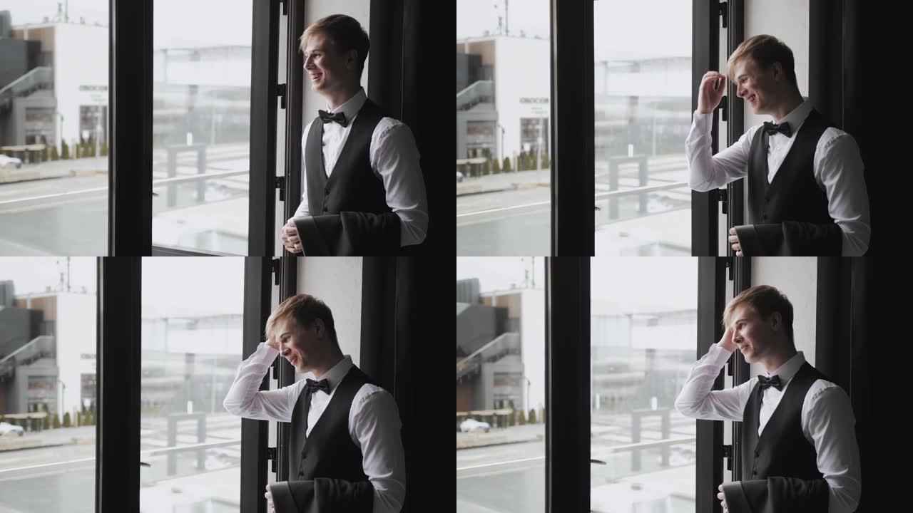靠近窗户的酒店房间里穿着黑色领结的新郎肖像。新郎快乐而微笑。婚礼当天。成功的商人，穿白衬衫的新郎。特