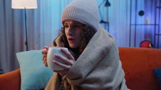生病的不健康女人戴着帽子裹着格子坐着，在沙发上喝热茶冻得瑟瑟发抖