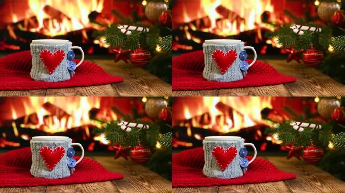 在燃烧的壁炉，红色格子和一棵圣诞树的背景下，在木制桌子上放着带有红色心形的蓝色针织罩子的热饮杯子