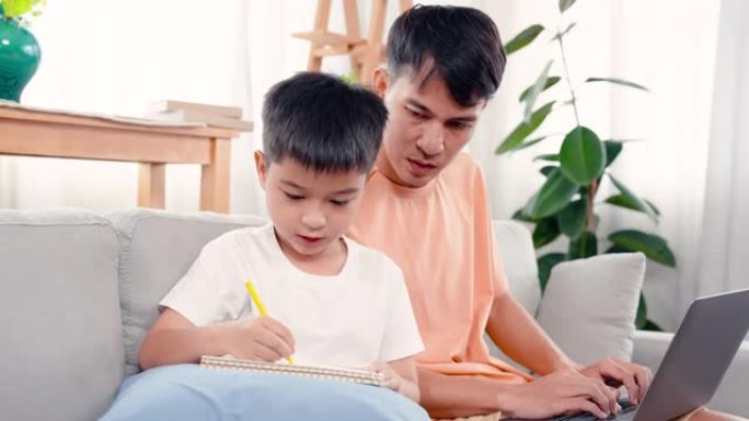 4K，一个亚洲单身父亲正坐在家里客厅的沙发上，教儿子做作业给老师，当时父亲正在自己的笔记本上工作。